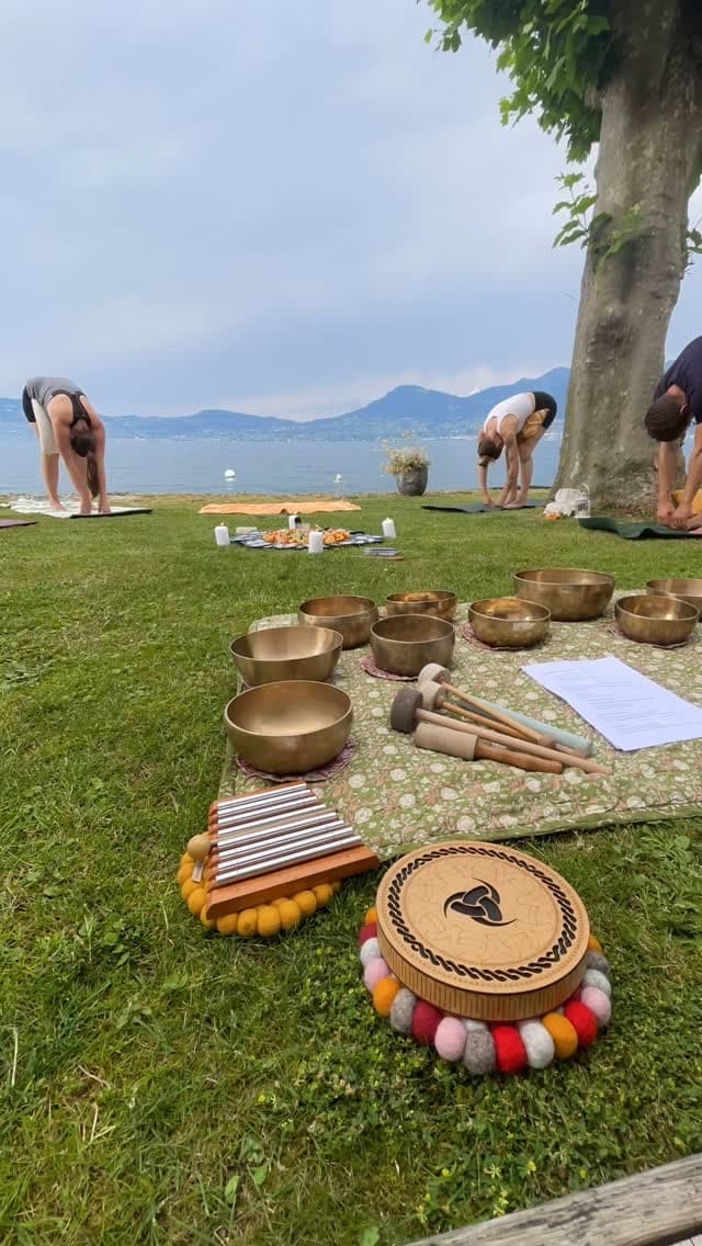 Des bols chantants, un carillon et un tambour posés sur une couverture dans l'herbe, au bord du lac, devant des personnes en posture