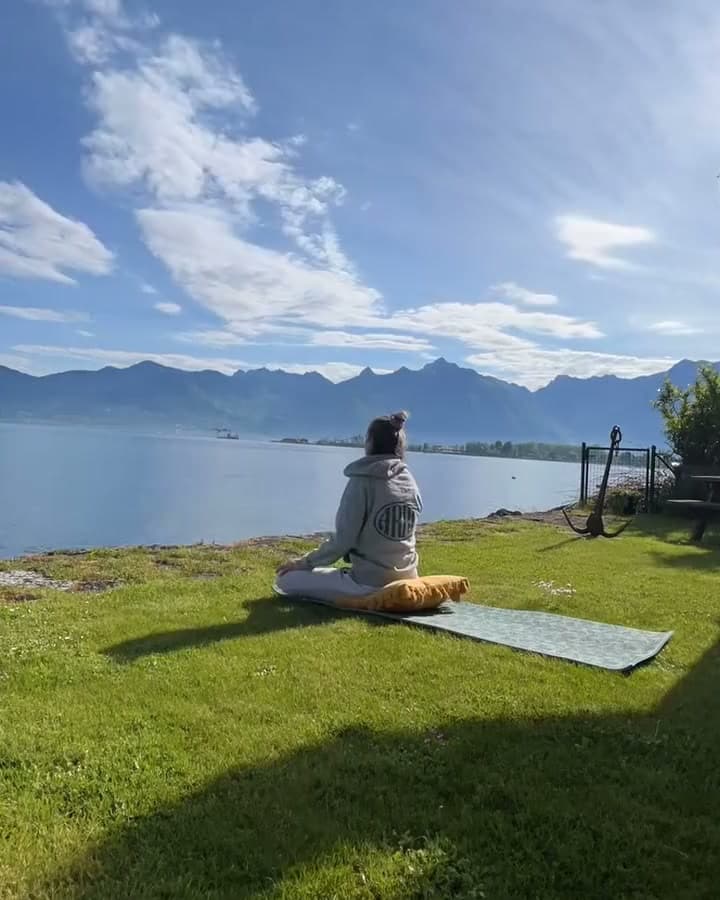 Léa assise en méditation sur la pelouse au bord du lac, face aux montagnes, pendant que passent les nuages