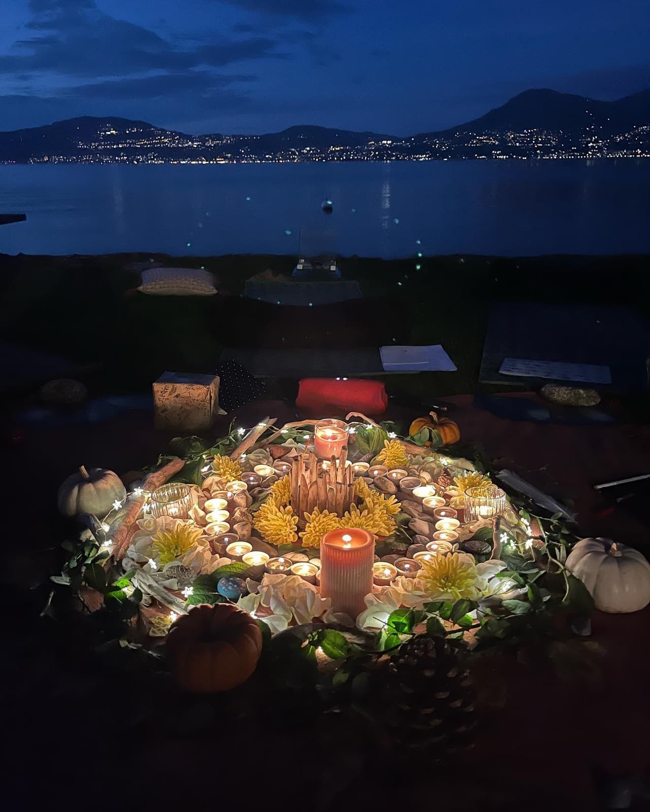 Un mandala de bougies, de fleurs jaunes et de petites courges, la nuit au bord du lac, face aux lumières de la rive